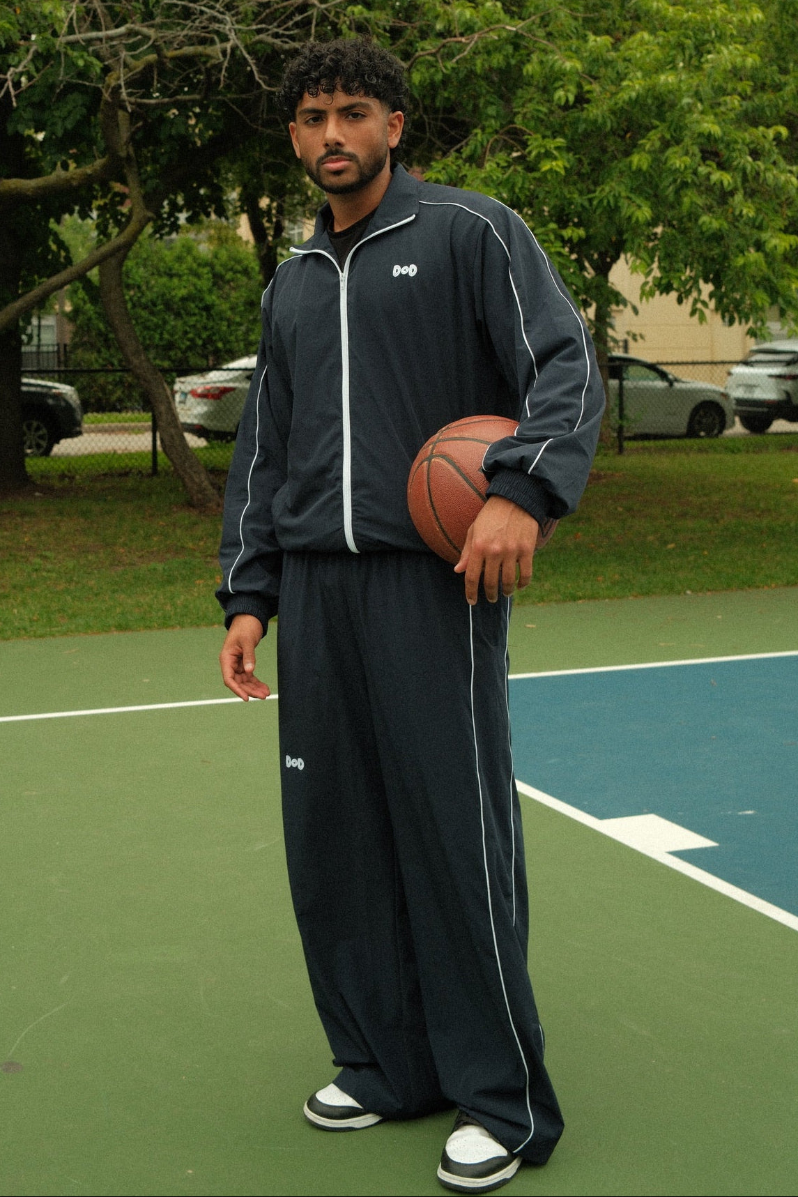 A man standing on an outdoor basketball court holding a basketball, wearing a dark navy blue tracksuit with white piping and a small white logo on the chest and thigh.